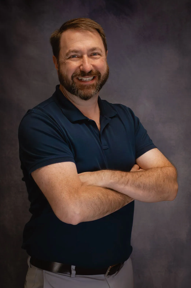 A man with brown hair and a beard wears a blue polo while crossing his arms and smiling in front of a gray background.