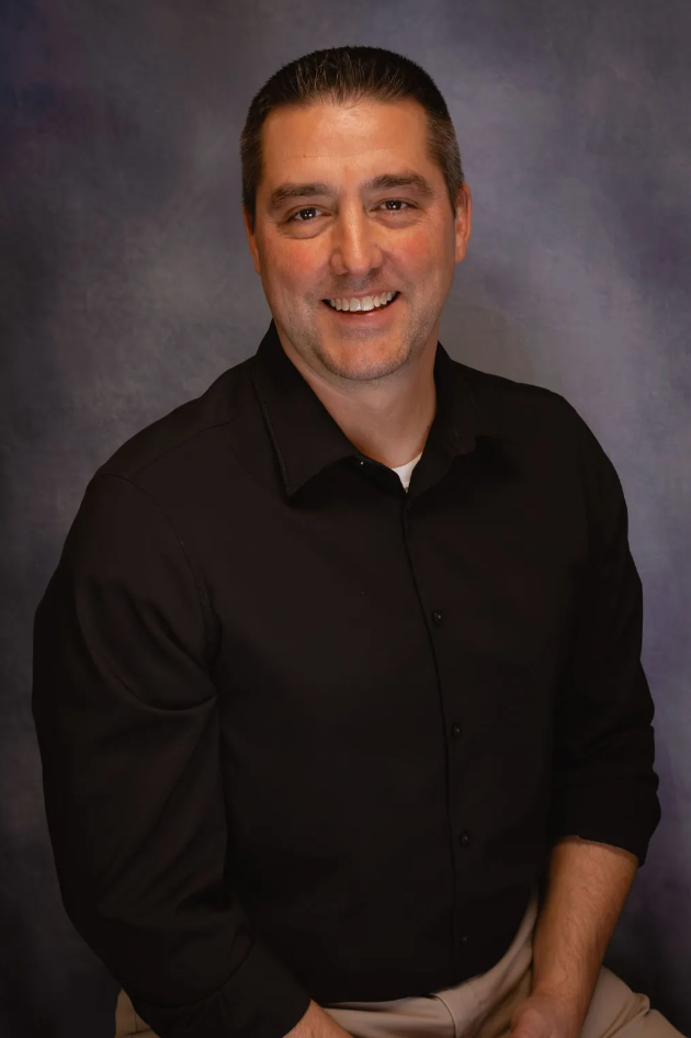 A man with dark brown hair and a black shirt smiles for a photo in front of a gray background.