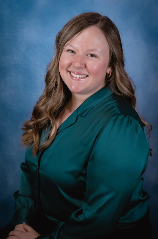 A women with long light brown hair and a teal blouse smiles in front of a blue background.