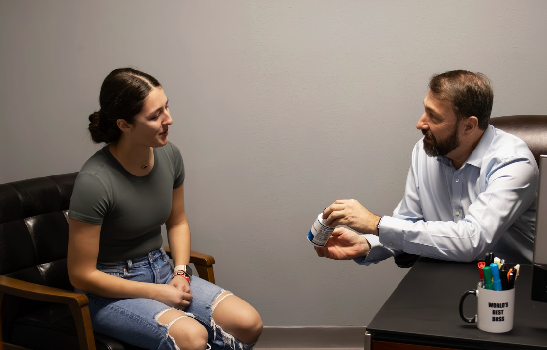 A male doctor presents a bottle of vitamins to a young women as they sit across from one another.